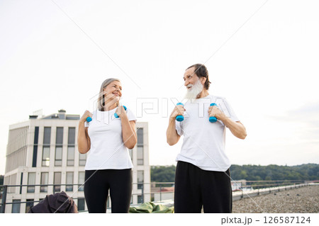 Elderly man and woman performing morning exercises with dumbbells outdoors on urban rooftop. Elderly man and woman performing morning exercises with dumbbells outdoors on urban rooftop. 126587124