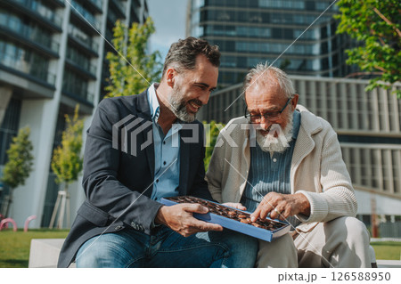 Senior dad and adult son sharing a box of sweets, chocolates on park bench. Senior dad and adult son sharing a box of sweets, chocolates on park bench. 126588950