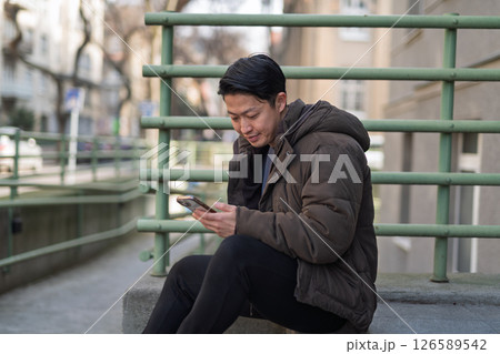 Young Asian man sitting on street ledge scrolling on smartphone 126589542