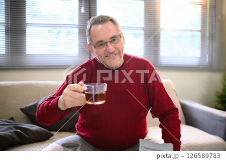 Happy retired man relaxing indoors with a glass cup of coffee in hand Happy retired man relaxing indoors with a glass cup of coffee in hand 126589783