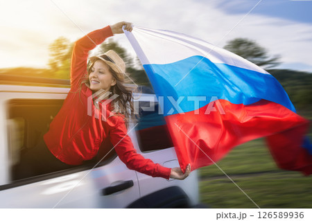 A woman joyfully waves the Russian flag from the window of a moving car as she embarks on a road trip to celebrate Russia Day. Dressed in a red outfit, she embodies the spirit of independence and 126589936