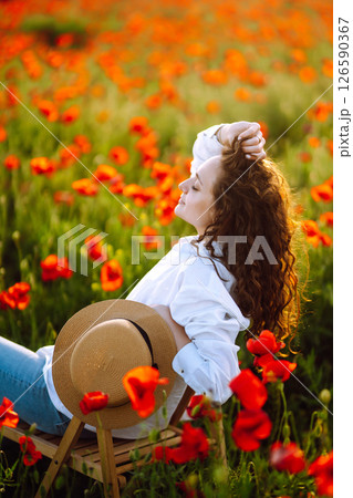 Young curly girl posing in a poppy field sitting on a chair. Summertime. 126590367