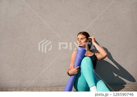 A young sportive woman enjoys a moment of relaxation after her yoga session, sitting against a wall while holding her mat. Girl exemplifies a fit and healthy lifestyle with a calm demeanor. 126590746