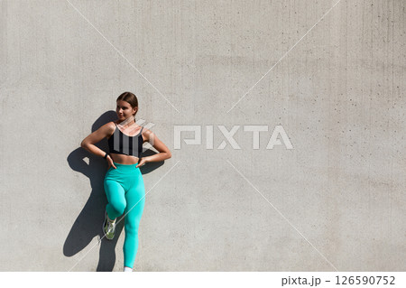 Confident young woman in athletic wear poses against a textured wall showcasing fitness and style, promoting an active lifestyle and wellness concept 126590752