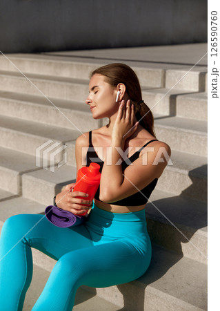 After an energizing workout, a young woman sits on the steps basking in the sun. Girl holds a water bottle in one hand and a yoga mat in the other, enjoying a moment of peace 126590760