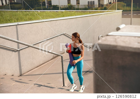 Young woman in activewear carrying yoga mat and drink, walking up outdoor stairs, showcasing fitness lifestyle and determination, with modern architecture in the background and bright daylight Young woman in activewear carrying yoga mat and drink, walking up outdoor stairs, showcasing fitness lifestyle and determination, with modern architecture in the background and bright daylight 126590812