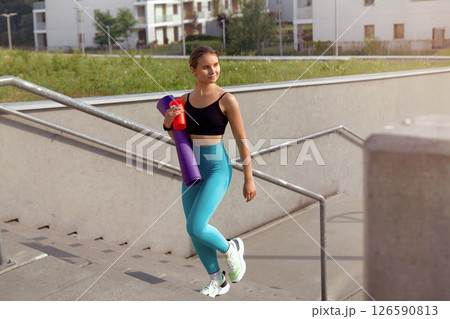 Young woman in athletic wear carrying yoga mat and water bottle, walking down outdoor stairs, surrounded by modern architecture and greenery, embodying a healthy lifestyle and fitness journey Young woman in athletic wear carrying yoga mat and water bottle, walking down outdoor stairs, surrounded by modern architecture and greenery, embodying a healthy lifestyle and fitness journey 126590813