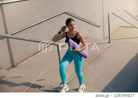 Young athletic woman carrying yoga mat while climbing outdoor stairs, dressed in vibrant activewear, showcasing fitness lifestyle and dedication to health and wellness in a modern urban environment 126590817