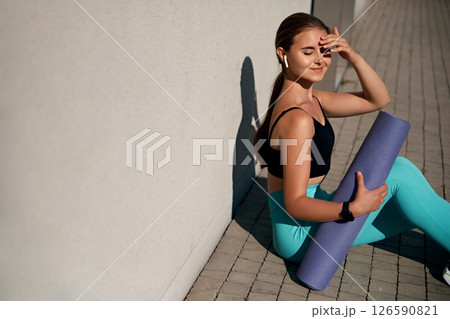 Young woman in athletic wear sitting on pavement, holding yoga mat, enjoying sunlight, with a relaxed expression, showcasing fitness lifestyle and mindfulness in an outdoor urban environment 126590821