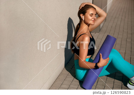 Young woman in athletic wear sitting on pavement with yoga mat, enjoying music through wireless earbuds, radiating calmness and focus in a serene outdoor environment, promoting wellness and fitness 126590823