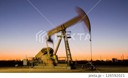 As dusk settles over the landscape an oil pump stands tall its arm moving rhythmically against the serene twilight. The horizon glows softly showcasing nature's beauty amidst industry. 126592021