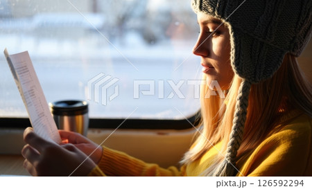 A young woman in a warm hat enjoys a quiet afternoon ride immersed in reading a menu as snow blankets the outside world. The sunlight casts a soft glow on her face adding warmth. 126592294