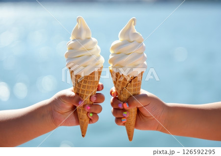 Two joyful hands hold up luscious ice cream cones glistening in the sunlight. The serene waterfront glimmers in the background evoking a sense of delightful summer fun. 126592295