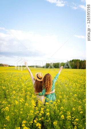 Two young women in beautiful dresses walk in a field with yellow blooming rapeseed. Concept of fun. Two young women in beautiful dresses walk in a field with yellow blooming rapeseed. Concept of fun. 126595598