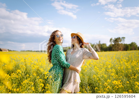Two young women in beautiful dresses walk in a field with yellow blooming rapeseed. Concept of fun. 126595604