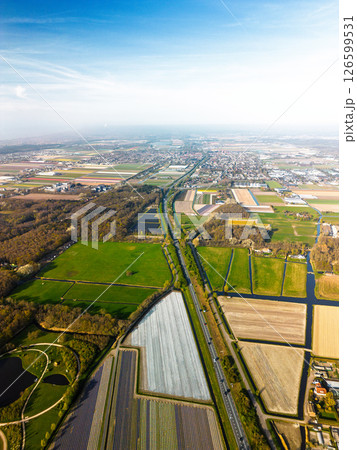 Aerial panoramic view of the Dutch countryside featuring farmland, green fields, forests, and a highway stretching through the landscape toward a distant town. Aerial panoramic view of the Dutch countryside featuring farmland, green fields, forests, and a highway stretching through the landscape toward a distant town. 126599531