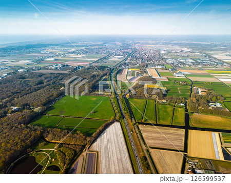 Aerial panoramic view of the Dutch countryside featuring farmland, green fields, forests, and a highway stretching through the landscape toward a distant town. Aerial panoramic view of the Dutch countryside featuring farmland, green fields, forests, and a highway stretching through the landscape toward a distant town. 126599537