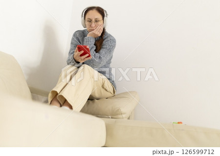Bored woman listening music with wireless headphones and browsing smartphone sitting on sofa 126599712