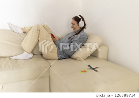 Woman relaxing on sofa listening to music and browsing smartphone Woman relaxing on sofa listening to music and browsing smartphone 126599714