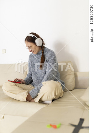 Woman wearing headphones listening to music and using smartphone on sofa at home 126599730