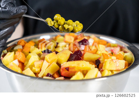 adding green peas to a homemade vinaigrette salad with beets, potatoes, carrots, and pickles in a metal bowl, chef in black gloves 126602525
