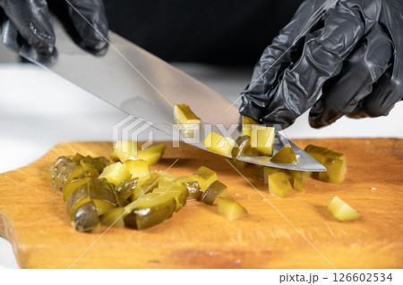 chopping pickles for olivier salad, close-up of a sharp knife cutting green cucumbers on a wooden board, hands in black gloves preparing ingredients chopping pickles for olivier salad, close-up of a sharp knife cutting green cucumbers on a wooden board, hands in black gloves preparing ingredients 126602534
