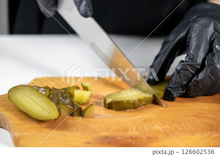 chef slicing pickles on a wooden board, hands in black gloves cutting a green cucumber with a sharp knife, preparing ingredients for a salad 126602536