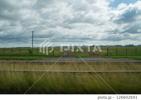 A Wide Grassy Field and Distant Airport Buildings Appear With Motion Blur Through a Tour Bus Window En Route to the Countryside Under a Cloudy Sky and Soft Natural Light. A Wide Grassy Field and Distant Airport Buildings Appear With Motion Blur Through a Tour Bus Window En Route to the Countryside Under a Cloudy Sky and Soft Natural Light. 126602691