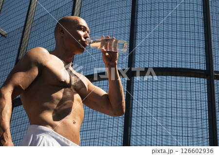 Athletes handsome sun-tanned shirtless man drinking water on outdoor sports ground. Concept water balance for hot season for active workout. Banner. Heat summer. Close up. Athletes handsome sun-tanned shirtless man drinking water on outdoor sports ground. Concept water balance for hot season for active workout. Banner. Heat summer. Close up. 126602936