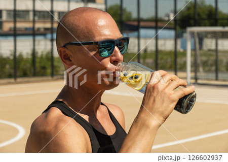 Athletes tanned man in black vest drinking water on outdoor sports ground. Water balance for heat season for active workout. Close up. Athletes tanned man in black vest drinking water on outdoor sports ground. Water balance for heat season for active workout. Close up. 126602937