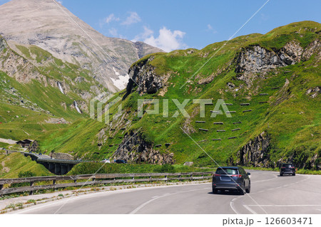 Cars drive Grossglockner High Alpine Road mountain green hills peaks blue sky background. Winding path through highland landscape offers scenic view and mountain driving experience 126603471