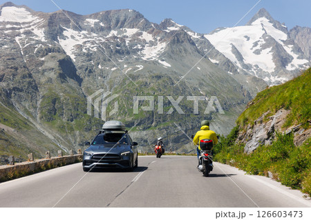 Cars and scooter drive Grossglockner High Alpine Road mountain green hills peaks blue sky background. Winding path through highland landscape offers scenic view and mountain driving experience 126603473