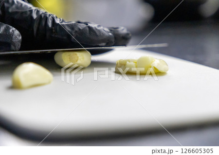 fresh garlic cloves being sliced on a white cutting board with sharp knife and gloved hand fresh garlic cloves being sliced on a white cutting board with sharp knife and gloved hand 126605305