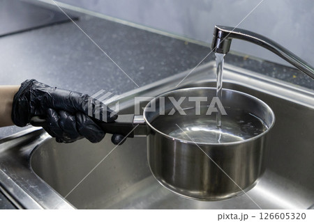 filling a stainless steel pot with water from a kitchen faucet, hand in black glove holding the handle over a sink 126605320