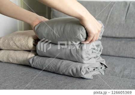 stacking gray linens on soft fabric sofa with hands carefully arranging folded bedding for tidy and organized home interior 126605369