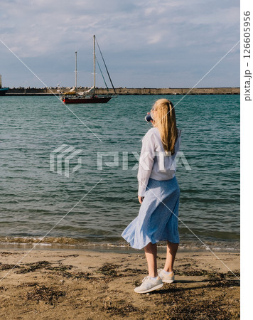 Blonde woman sipping coffee on beach, enjoying view of red sailboat gliding away on sunny day, framed by backdrop of fluffy clouds and endless blue sky. Blonde woman drinks coffee, red sailboat, ocean Blonde woman sipping coffee on beach, enjoying view of red sailboat gliding away on sunny day, framed by backdrop of fluffy clouds and endless blue sky. Blonde woman drinks coffee, red sailboat, ocean 126605956