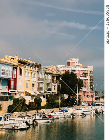 Colorful buildings stand along marina with moored boats under cloudy sky, creating vibrant and picturesque scene in Spanish coastal town. Colorful marina buildings lining waterfront with moored boats Colorful buildings stand along marina with moored boats under cloudy sky, creating vibrant and picturesque scene in Spanish coastal town. Colorful marina buildings lining waterfront with moored boats 126605958