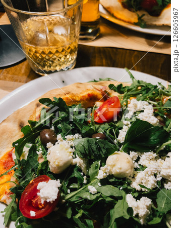 Close up of pizza topped with fresh arugula, juicy tomatoes, salty olives, crumbled feta, and creamy mozzarella on white plate, paired with refreshing alcoholic beverages. Beer, rustic white plate 126605967