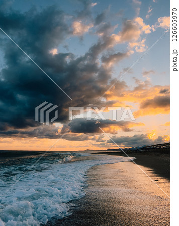 Ocean waves crashing on sandy beach during dramatic sunset illuminating clouds with orange and blue hues, creating picturesque seascape. Powerful ocean waves breaking against sandy coastline Ocean waves crashing on sandy beach during dramatic sunset illuminating clouds with orange and blue hues, creating picturesque seascape. Powerful ocean waves breaking against sandy coastline 126605970