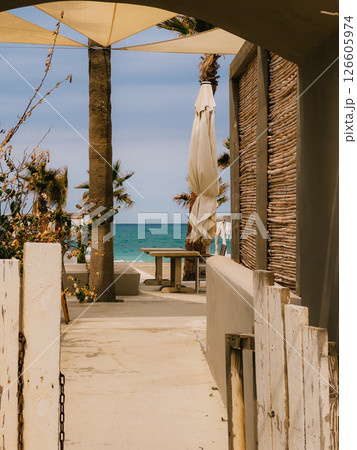 View of beach club entrance with sunshade, palm trees, table, wooden fence and turquoise sea in background, inviting to relaxation and summer vibes. Wooden fence framing luxurious beach club entrance View of beach club entrance with sunshade, palm trees, table, wooden fence and turquoise sea in background, inviting to relaxation and summer vibes. Wooden fence framing luxurious beach club entrance 126605974