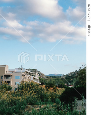 Lush green vegetation partially obscures modern hotel against rolling hills under dynamic, cloud filled sky, creating tranquil and inviting scene. Verdant landscape, undulating terrain, cloudscape Lush green vegetation partially obscures modern hotel against rolling hills under dynamic, cloud filled sky, creating tranquil and inviting scene. Verdant landscape, undulating terrain, cloudscape 126605976