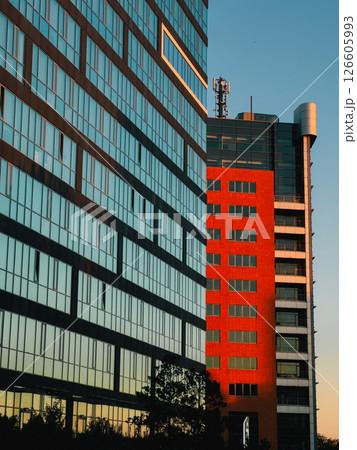 Glass and red brick facades of modern office buildings reflecting warm sunset light, creating a vibrant contrast against the clear blue sky. Glass facades and red brick walls reflecting golden sunset 126605993