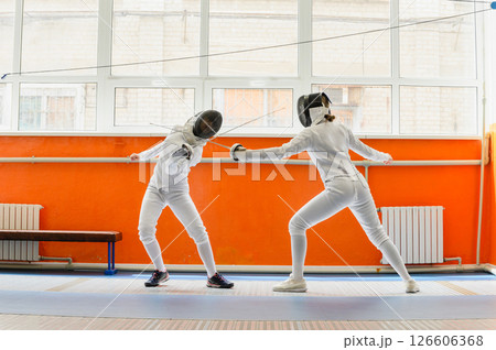 Female fencer striking opponent in bright indoor fencing hall. Female fencer striking opponent in bright indoor fencing hall. 126606368