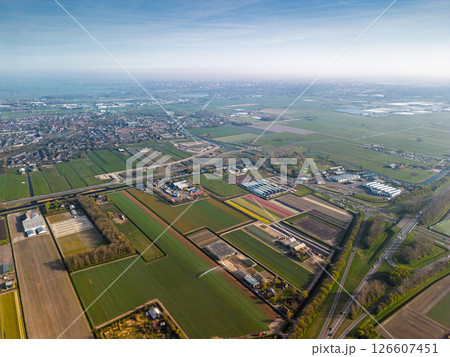 Aerial panoramic view of the Dutch countryside featuring farmland, green fields, forests, and a highway stretching through the landscape toward a distant town. Aerial panoramic view of the Dutch countryside featuring farmland, green fields, forests, and a highway stretching through the landscape toward a distant town. 126607451