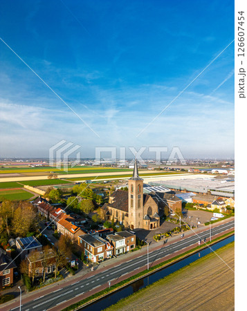 Aerial view of a picturesque village church with a tall steeple, surrounded by houses and farmland under a bright blue sky in a serene countryside setting. 126607454