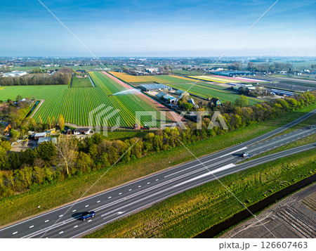 Aerial panoramic view of the Dutch countryside featuring farmland, green fields, forests, and a highway stretching through the landscape toward a distant town. Aerial panoramic view of the Dutch countryside featuring farmland, green fields, forests, and a highway stretching through the landscape toward a distant town. 126607463