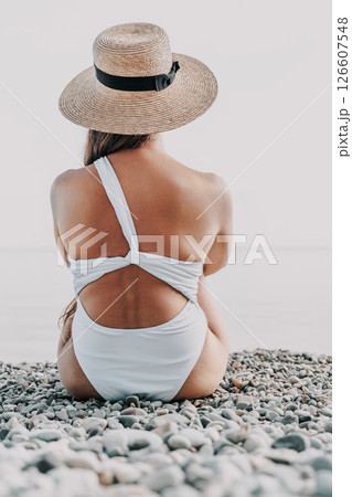 A woman is sitting on a beach wearing a straw hat and a white bikini. The beach is rocky and the sky is blue. 126607548