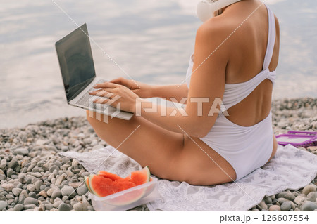 A woman is sitting on the beach with a laptop open in front of her. She is wearing a white bikini and has a pink headband on. Concept of relaxation and leisure. 126607554