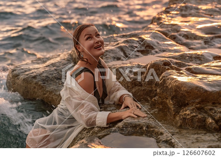 Woman Beach Rocks Sunset - A woman in a white shirt and black swimsuit smiles while sitting on rocks by the ocean at sunset. 126607602