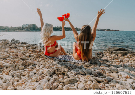 Watermelon Beach Summer: Two women celebrate summer joy, enjoying watermelon slices on a rocky beach. 126607613